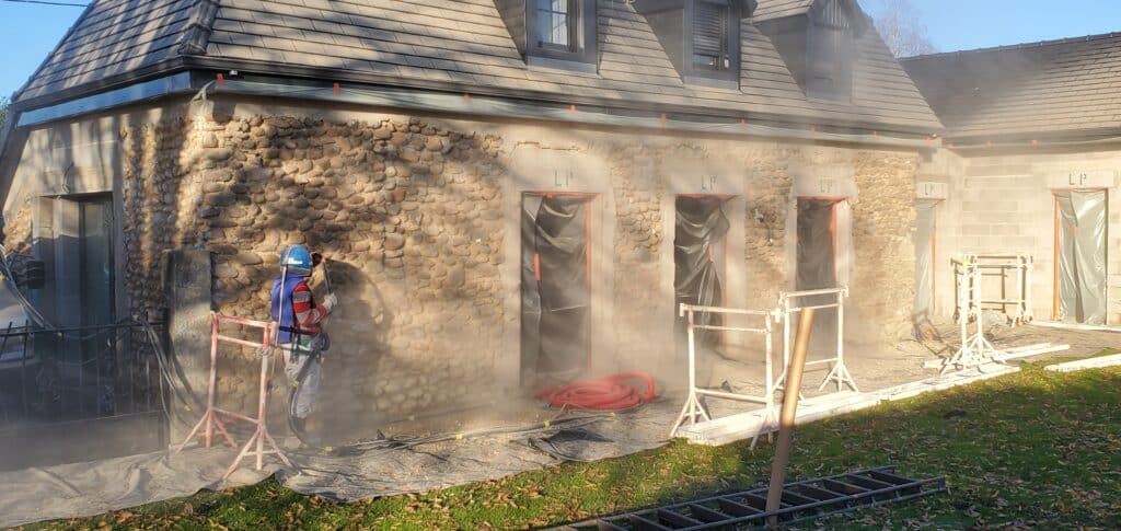 Ouvrier en combinaison protectrice nettoyant une façade en pierre et crépi d'un bâtiment sous la poussière. Ouvertures masquées, texte 'LP' visible.