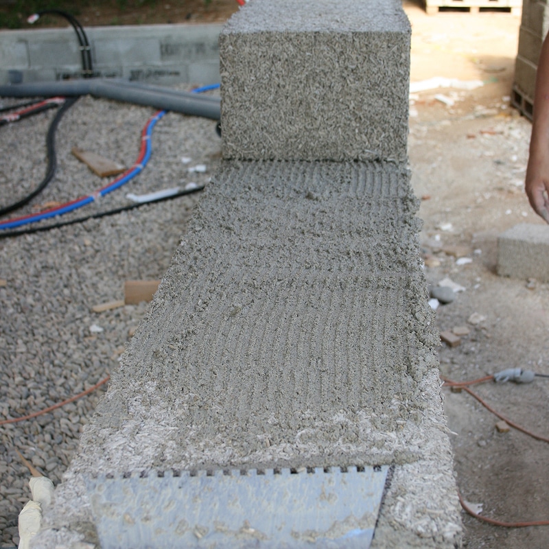 Maçon appliquant du mortier cranté sur un bloc de construction gris. Une truelle dentelée repose sur le bloc texturé. Gravier et tuyaux en arrière-plan.