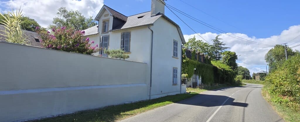 Maison crème datée 1892 aux volets bleus, mur blanc et arbustes fleuris le long d'une route de campagne ensoleillée.