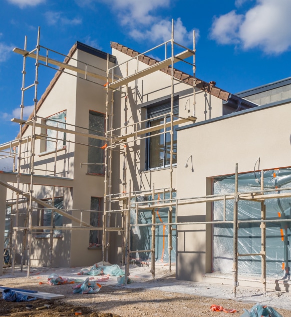 Maison neuve en construction avec échafaudages, façade beige, fenêtres protégées, sous un ciel bleu. Travaux de rénovation.