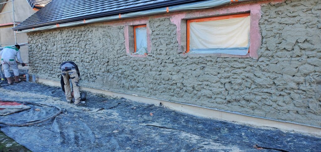 Deux ouvriers appliquent un enduit texturé sur la façade d'une maison en rénovation. Les fenêtres sont protégées par du film.