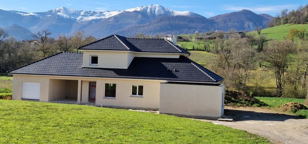 Maison neuve beige sur herbe verte, toit foncé. Arrière-plan de montagnes enneigées et ciel bleu. Paysage lumineux de printemps.