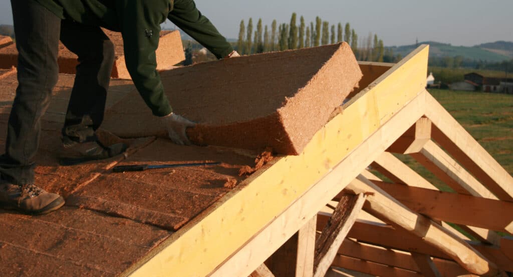 Ouvrier installant des panneaux d'isolation en fibre naturelle sur une toiture en bois. Lumière dorée sur chantier rural.
