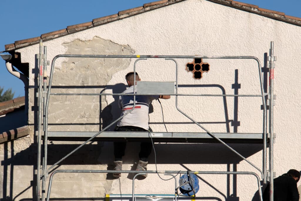 Ouvrier sur échafaudage rénovant la façade crépie d'une maison, enduit écaillé. Ombre, tuiles et gouttière. Poteau "ALTRAS".