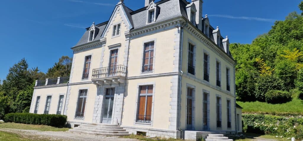 Élégant manoir jaune pâle et pierre grise, toit en ardoise, balcons en fer forgé, escalier courbé, au milieu d'un jardin luxuriant sous ciel bleu.
