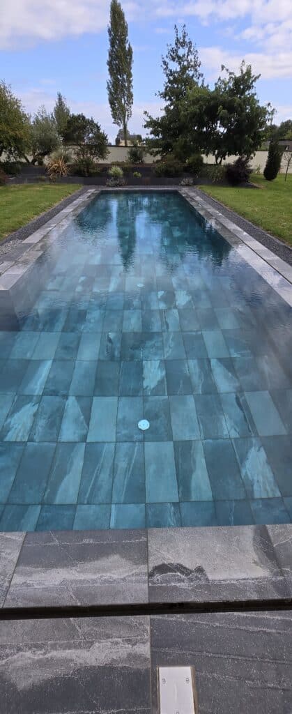 Piscine rectangulaire moderne avec carrelage bleu-vert et margelles sombres. Jardin paysager luxuriant, ciel bleu nuageux reflété.