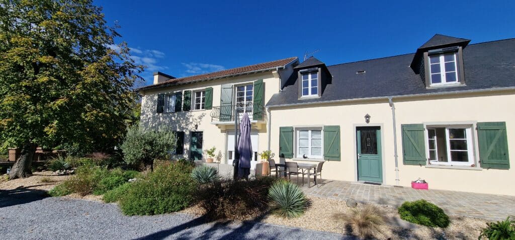 Maison Campagne avec Volets Verts et Jardin Ensoleillé Maison de campagne claire avec volets verts, terrasse pavée, jardin de gravier et grand arbre feuillu sous un ciel bleu intense.